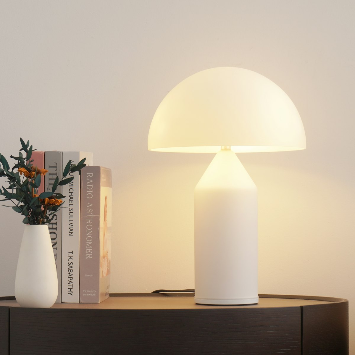 A round white table lamp glowing on a sideboard beside books and a vase